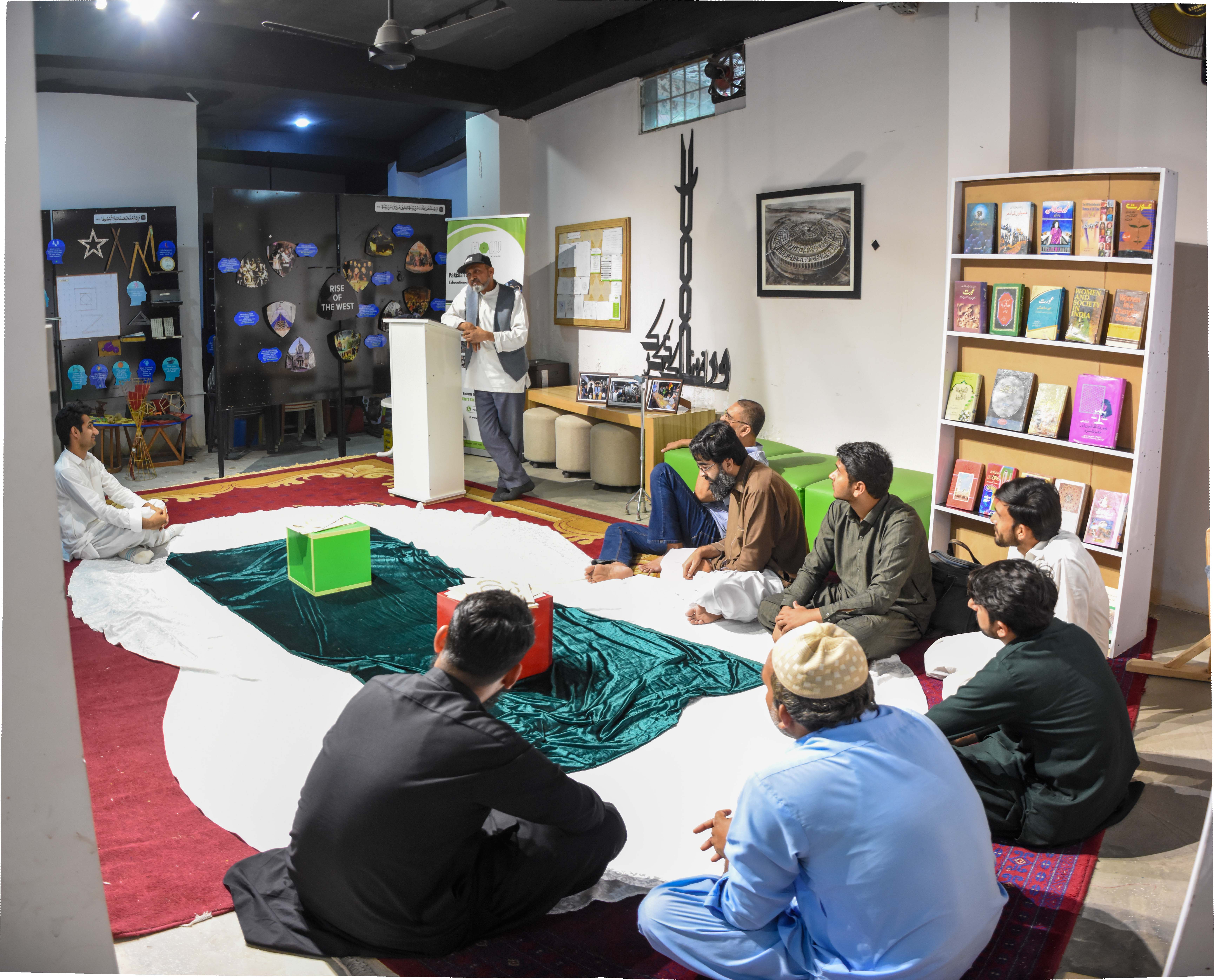 Guidance — A speaker addresses a small circle of men and boys seated on the carpet near the bookshelves, opening the gathering with words of guidance.